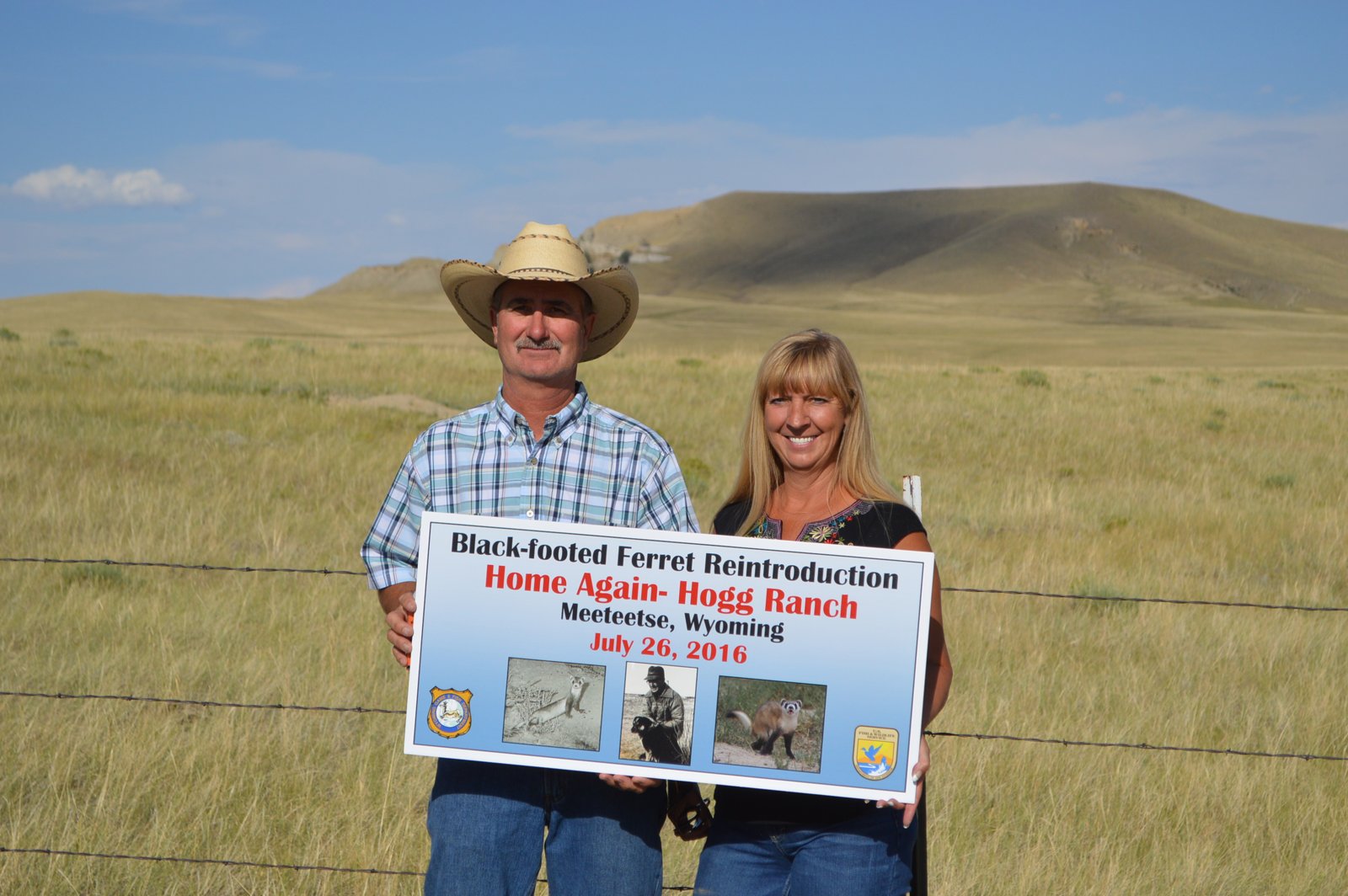 A man in a cowboy hat and a woman stand by a barbed wire fence on a Wyoming grassland holding a sign reading Black-footed Ferret Reintroduction, Home Again, Hogg Ranch, Meeteetse, Wyoming, July 26, 2016