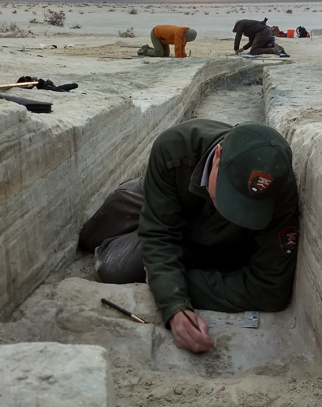 A researcher lies in a trench gently brushing sediment from a fossil footprint while two colleagues work behind him